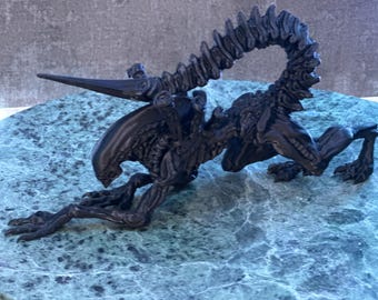 May include: A black alien creature figurine with a long, sharp tail and a pointed head. The figurine is posed in a crouching position, with its head lowered and its tail curled around its body.