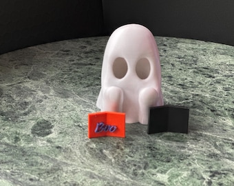May include: A white ghost figurine with two oval eye openings, positioned behind a small orange rectangular sign with the word Boo written in purple script and a separate small black rectangular sign folded at a right angle.