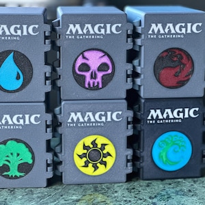 May include: Six grey Magic: The Gathering card storage boxes. Each box features a different symbol in a circle, including a blue water drop, a purple skull, and a red symbol.