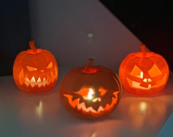 May include: Three orange pumpkin-shaped decorations with carved, illuminated faces. Each pumpkin features a unique, jagged expression with glowing light emanating from within, creating a warm, bright orange light against the dark background.
