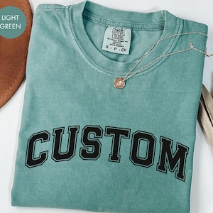 以下が含まれることがあります： 黒いブロック体の「CUSTOM」という文字が入った、明るい緑色のTシャツ。Tシャツはクルーネックで半袖です。ペンダント付きのシルバーのネックレスがTシャツの上にかけられています。