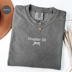 Peut inclure: Un t-shirt gris Comfort Colors avec le texte "chapter 30" et un motif de nœud. Un collier argenté avec un pendentif rond est posé sur le t-shirt. Le t-shirt est plié sur une surface blanche.