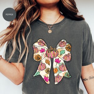 以下が含まれることがあります： カボチャと花柄の大きなリボンデザインが特徴の濃いグレーのTシャツ。リボンは、ピンク、オレンジ、緑の色合いのカボチャ、花、葉が描かれたクリーム色の背景です。「Pepper」の文字が左上に丸で囲まれています。