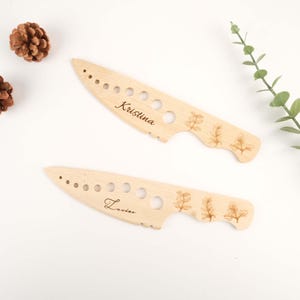 May include: Two wooden cheese knives with personalised names "Kristina" and "Louise" engraved on them. Each knife has a decorative design of a sprig of herbs.