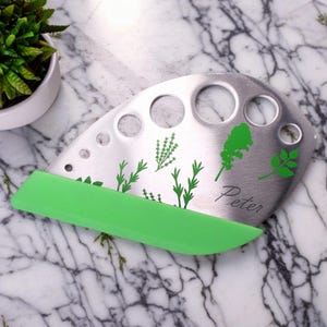 May include: A stainless steel herb chopper with a green handle. The chopper has multiple round holes and a design of herbs on the surface. The word "Peter" is engraved on the chopper.
