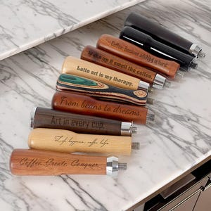 May include: A set of eight wooden espresso tamper handles with different wood grains and engraved sayings. The sayings include "Coffee. Because adulting is hard." "Wake up & smell the coffee." "Latte art is my therapy." "From beans to dreams." "Art in every cup." "Life happens, coffee helps." "Coffee. Create. Conquer."