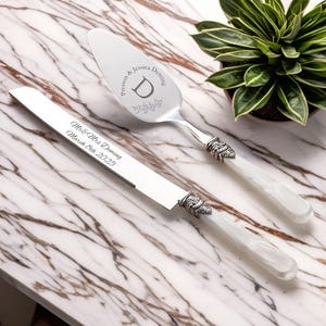 May include: A silver cake knife and server set with white handles. The knife has the text "Mr & Mrs Deming March 8th 2025". The server has the text "Preston & Jessica Deming" and a decorative "D". The set is on a marble surface.