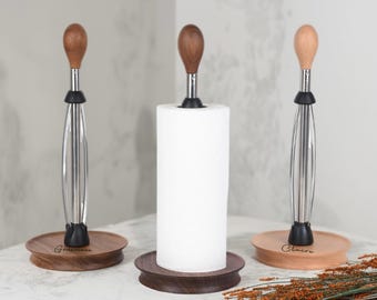 Puede incluir: Tres portarrollos de papel de cocina con bases y tapas de madera. Los soportes tienen un centro de acero inoxidable y un anillo de goma negro. Las bases están grabadas con nombres. Un rollo de papel de cocina blanco está en un soporte.