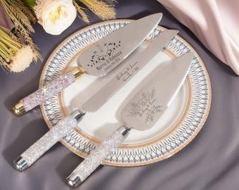 May include: A set of three silver cake serving utensils with crystal-studded handles. The server and knife are engraved with names and dates. The set is displayed on a decorative plate, with floral accents and a soft background.
