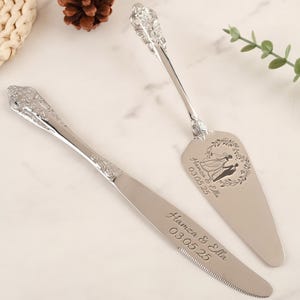 Könnte beinhalten: Ein silbernes Kuchenmesser- und -Server-Set mit verzierten Griffen. Der Server hat ein herzförmiges Design mit einer Silhouette eines Brautpaares im Inneren. Messer und Server sind mit den Namen "Hamza & Ella" und dem Datum "03.05.25" graviert.