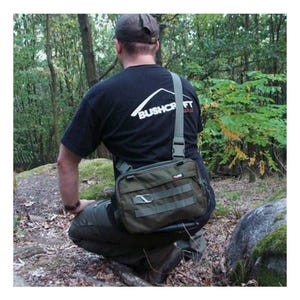 Könnte beinhalten: Olivgrüne Umhängetasche mit mehreren Taschen und Riemen. Die Tasche wird quer über dem Körper getragen. Die Person auf dem Bild trägt ein schwarzes T-Shirt mit dem weißen Text "Bushcraft". Der Hintergrund ist eine Waldlandschaft.