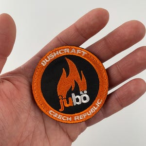 Può includere: Toppa rotonda arancione e nera con ricamo bianco. La toppa presenta un disegno a fiamma con il testo "Bushcraft Jubo Repubblica Ceca".