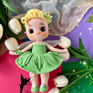 Puede incluir: Una muñeca de hada de crochet con cabello rubio, vestido verde y alas blancas. La muñeca está de pie sobre una superficie rosa y verde con tulipes blancas.