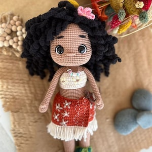 May include: Crocheted doll with brown skin, black curly hair, and a flower in her hair. She is wearing a brown and white crocheted dress with a flower pattern. The doll is sitting on a brown surface.