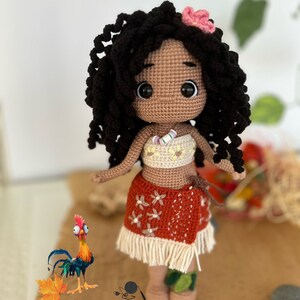May include: A crocheted doll with brown skin, black curly hair, and a pink flower in her hair. She is wearing a white and yellow top, a red and white skirt with a white fringe, and a white necklace. She is standing on a brown surface with a rooster and some leaves.