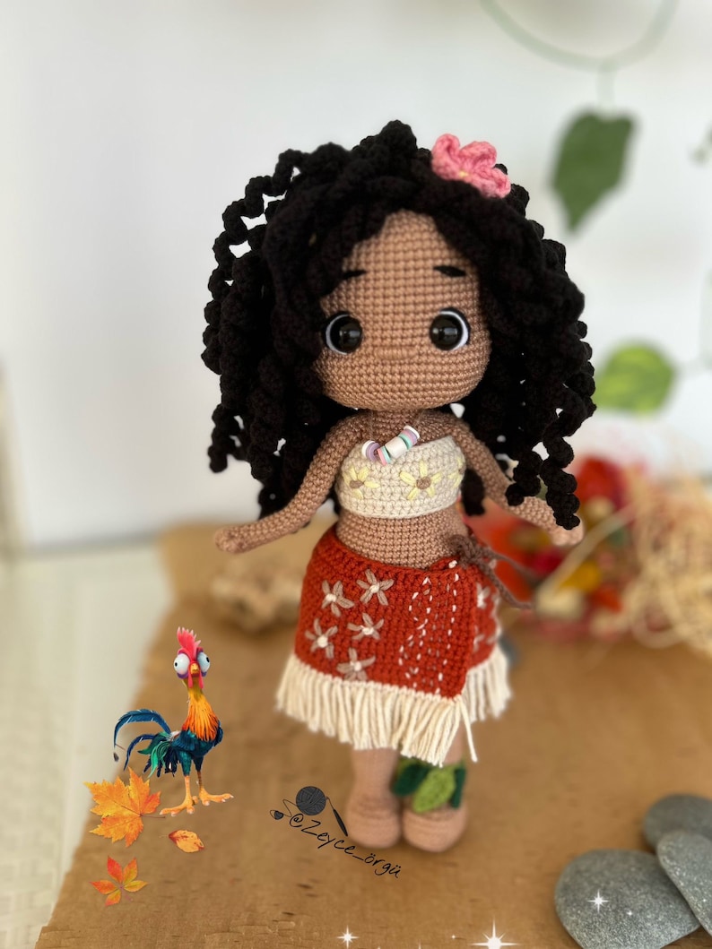May include: A crocheted doll with brown skin, black curly hair, and a pink flower in her hair. She is wearing a white and yellow top, a red and white skirt with a white fringe, and a white necklace. She is standing on a brown surface with a rooster and some leaves.