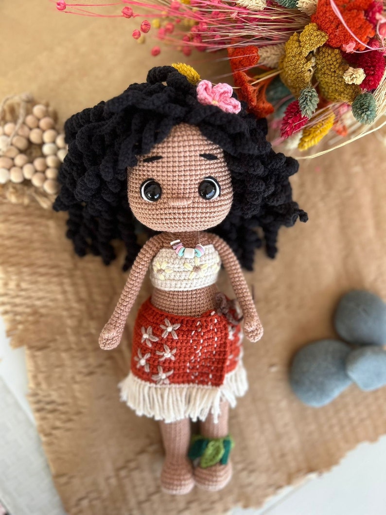 May include: Crocheted doll with brown skin, black curly hair, and a flower in her hair. She is wearing a brown and white crocheted dress with a flower pattern. The doll is sitting on a brown surface.