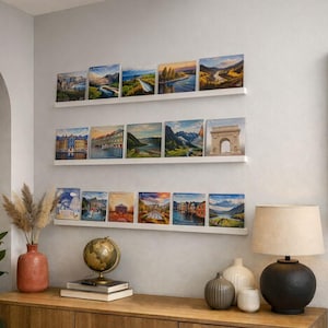 Op de afbeelding: Een display van ingelijste landschapskunstafdrukken, gerangschikt op witte planken tegen een lichtgrijze muur. De afdrukken tonen verschillende schilderachtige locaties, waaronder bergen, rivieren en architectonische bezienswaardigheden. Een houten kast met decoratieve items staat eronder.