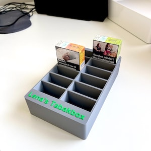 Könnte beinhalten: Eine graue Plastikbox mit Fächern zum Aufbewahren von Zigaretten. Die Box hat den Text "Lena's Tabakbox" in grünen Buchstaben auf der Vorderseite. Zwei Zigarettenpackungen sind in der Box sichtbar, eine mit einem Bild eines Babys und dem Text "Rauchen verursacht 9 von 10 Lungenkarzinomen" und die andere mit einem Bild einer Frau und dem Text "Rauchen mindert Ihre Fruchtbarkeit."
