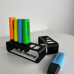 May include: A black plastic holder with four slots for highlighters. The holder has the words "Your Name" printed on it. There are four highlighters in the holder: orange, yellow, blue, and green. The highlighters are all Stabilo Boss Original brand. There is also a blue Stabilo Boss Original highlighter lying on a white surface.