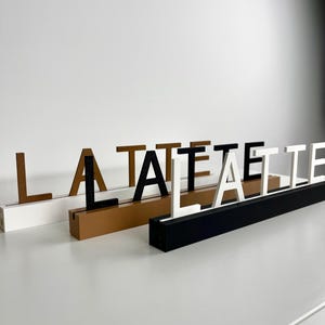 May include: Three rectangular bases in white, tan, and black, each featuring a slot holding upright letters. The white base displays brown letters, the tan base holds black letters, and the black base features white letters spelling out LA LATTE.