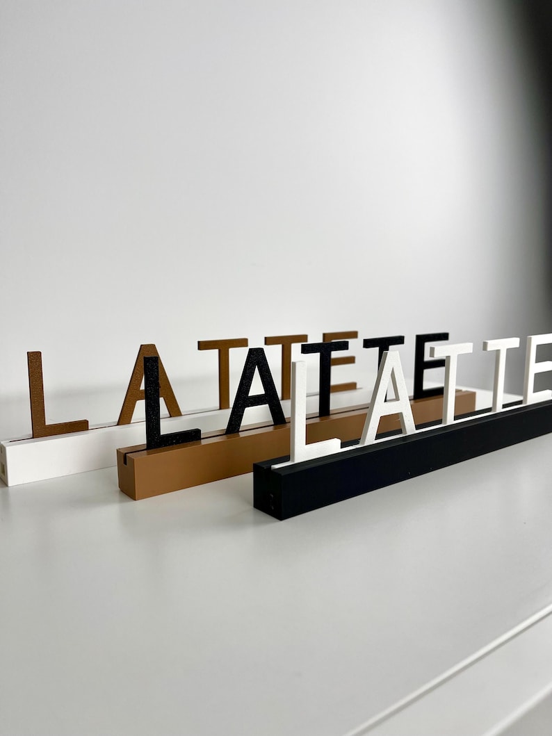 May include: Three rectangular bases in white, tan, and black, each featuring a slot holding upright letters. The white base displays brown letters, the tan base holds black letters, and the black base features white letters spelling out LA LATTE.