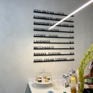 May include: A wall-mounted menu board with black horizontal rails displaying items including Iced Matcha Latte, Strawberry/Mango, Raspberry White Choc, Coconut Cloud, Lychee Cloud, Lavender, Matchagato, Banana Pudding, and Xoxo Cream Topping.