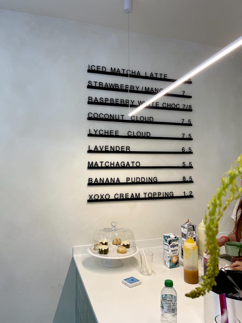May include: A wall-mounted menu board with black horizontal rails displaying items including Iced Matcha Latte, Strawberry/Mango, Raspberry White Choc, Coconut Cloud, Lychee Cloud, Lavender, Matchagato, Banana Pudding, and Xoxo Cream Topping.