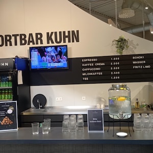 May include: A Sportbar Kuhn counter featuring a black menu board with white text listing beverages and prices, a glass beverage dispenser, a glass cake dome with pastries, a small refrigerator with bottled drinks, and a coffee machine.