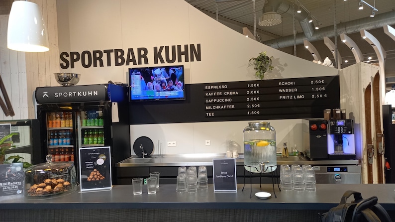 May include: A Sportbar Kuhn counter featuring a black menu board with white text listing beverages and prices, a glass beverage dispenser, a glass cake dome with pastries, a small refrigerator with bottled drinks, and a coffee machine.