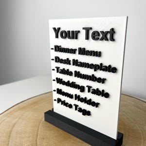May include: A white rectangular sign with black text that reads "Your Text" and lists options like "Dinner Menu" and "Desk Nameplate." The sign is held by a black rectangular base, suitable for various display purposes.