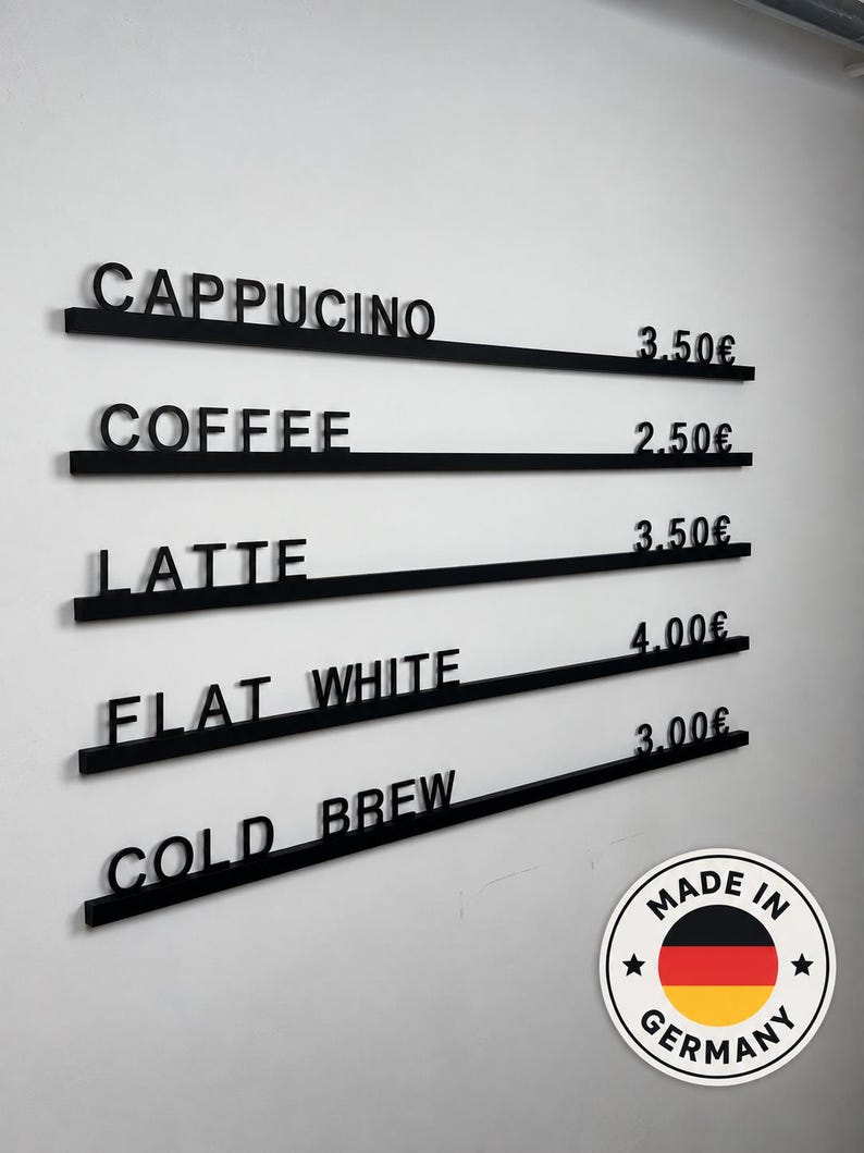 May include: A wall-mounted menu board featuring five horizontal black rails with raised black lettering and prices. The text reads CAPPUCINO 3.50&euro;, COFFEE 2.50&euro;, LATTE 3.50&euro;, FLAT WHITE 4.00&euro;, and COLD BREW 3.00&euro;. A circular Made in Germany sticker is visible.