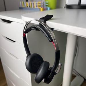 May include: Black headphones with a silver headband and red accents are hanging on a desk using a black desk clip.