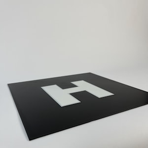 May include: A square, black sign with a white letter "H" in the center. The sign is set against a plain white background. The image is well-lit, highlighting the contrast between the black and white elements.