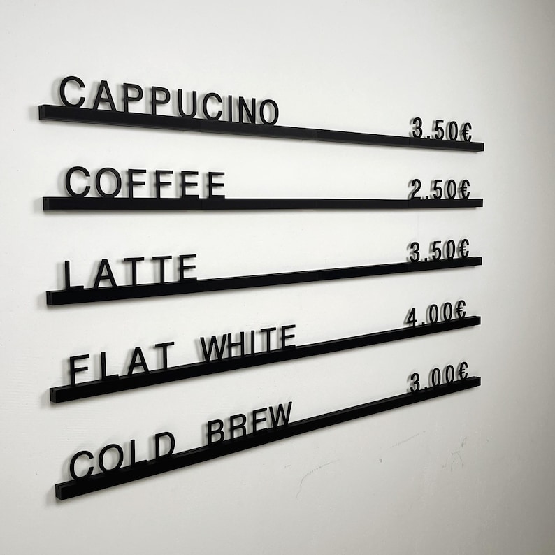 May include: A wall-mounted menu board featuring five horizontal black rails with individual black lettering and pricing. The text reads: CAPPUCINO 3.50&euro;, COFFEE 2.50&euro;, LATTE 3.50&euro;, FLAT WHITE 4.00&euro;, and COLD BREW 3.00&euro;, displayed in a clean, modern font.