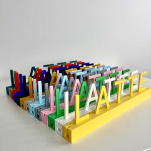 May include: A collection of multi-colored 3D printed letter stands arranged in rows. Each stand features a single upright letter attached to a rectangular base. The letters appear in various colors including yellow, white, green, pink, blue, and black.