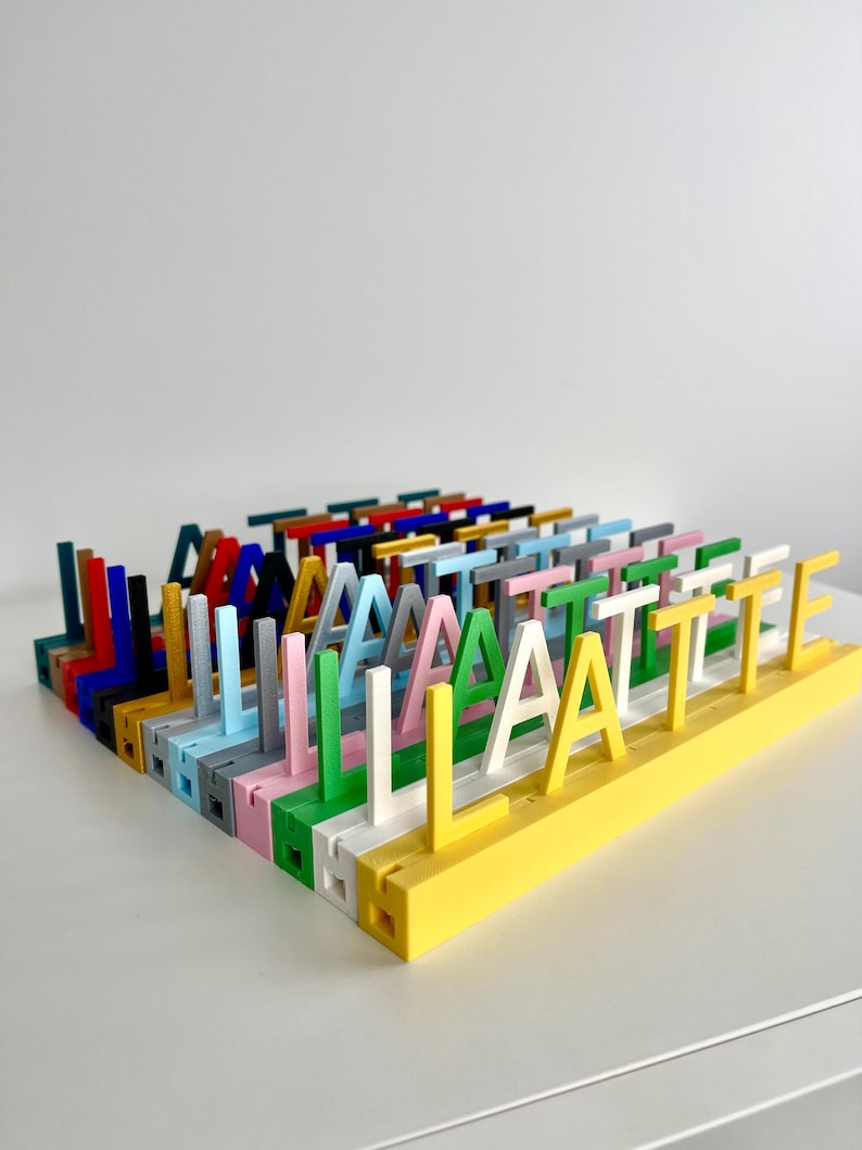 May include: A collection of multi-colored 3D printed letter stands arranged in rows. Each stand features a single upright letter attached to a rectangular base. The letters appear in various colors including yellow, white, green, pink, blue, and black.