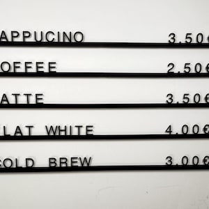May include: A black wall-mounted menu board featuring five horizontal rails with individual black lettering and pricing. The text reads: CAPPUCINO 3.50&euro;, COFFEE 2.50&euro;, LATTE 3.50&euro;, FLAT WHITE 4.00&euro;, and COLD BREW 3.00&euro;, displayed in a clean, sans-serif font.