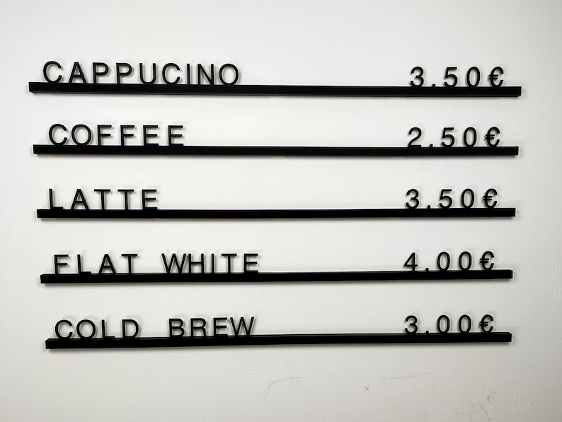 May include: A black wall-mounted menu board featuring five horizontal rails with individual black lettering and pricing. The text reads: CAPPUCINO 3.50&euro;, COFFEE 2.50&euro;, LATTE 3.50&euro;, FLAT WHITE 4.00&euro;, and COLD BREW 3.00&euro;, displayed in a clean, sans-serif font.