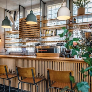 May include: A coffee bar interior featuring a wooden slatted counter with three stools, a stainless steel La Marzocco espresso machine, and a menu board listing coffee drinks. Three pendant lights hang above, with shelves displaying glassware, bottles, and plants.