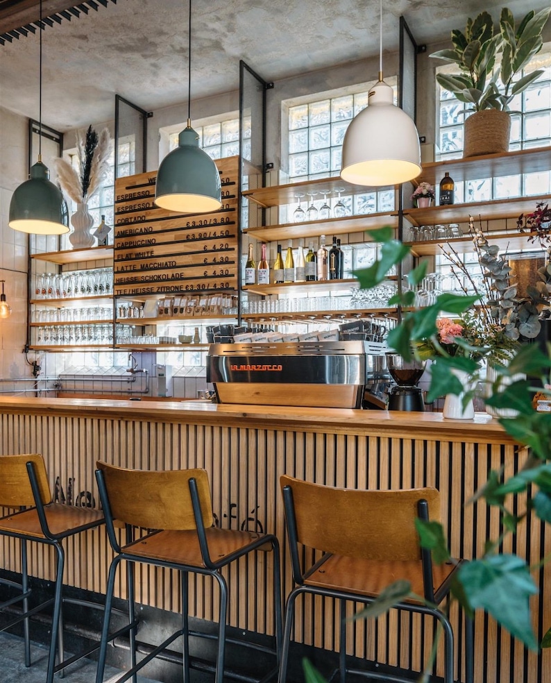 May include: A coffee bar interior featuring a wooden slatted counter with three stools, a stainless steel La Marzocco espresso machine, and a menu board listing coffee drinks. Three pendant lights hang above, with shelves displaying glassware, bottles, and plants.