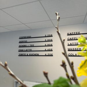 May include: A wall-mounted menu board with black horizontal rails displaying beverage and food items in white text. The menu lists Espresso 2.20&euro;, Doppio 3.80&euro;, Schoki / Tee 3.90&euro;, Chai Latte 4.40&euro;, and Torte & Kuchen 4.20&euro;, with additional items partially visible.