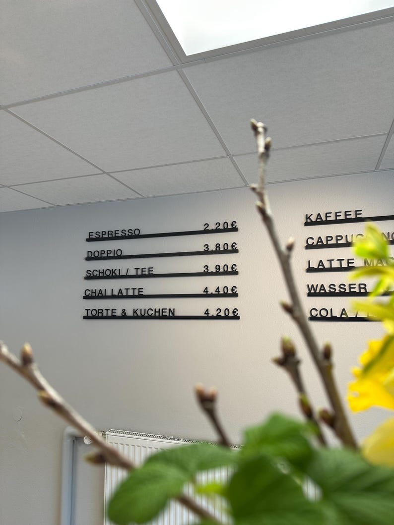 May include: A wall-mounted menu board with black horizontal rails displaying beverage and food items in white text. The menu lists Espresso 2.20&euro;, Doppio 3.80&euro;, Schoki / Tee 3.90&euro;, Chai Latte 4.40&euro;, and Torte & Kuchen 4.20&euro;, with additional items partially visible.