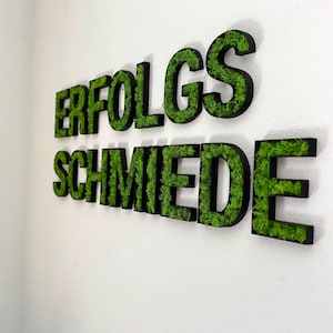 Pu&ograve; includere: Insegna a parete con la scritta "ERFOLGSSCHMIEDE" in grandi lettere tridimensionali. Le lettere hanno un bordo nero e sono riempite di muschio verde, creando un aspetto naturale e strutturato su una parete bianca.