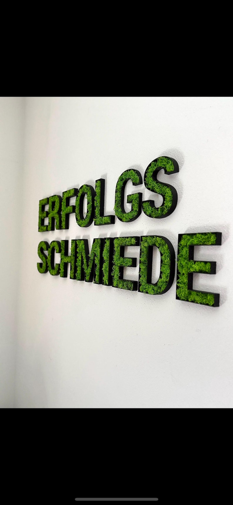 Pu&ograve; includere: Insegna a parete con la scritta "ERFOLGSSCHMIEDE" in grandi lettere tridimensionali. Le lettere hanno un bordo nero e sono riempite di muschio verde, creando un aspetto naturale e strutturato su una parete bianca.