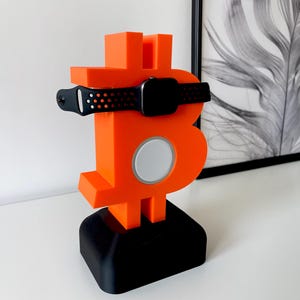 Pode incluir: Símbolo Bitcoin laranja impresso em 3D com uma pulseira de relógio preta e uma base de carregamento preta.