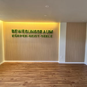 Pu&ograve; includere: Una stanza vuota con pavimento in legno e pareti chiare. La parete di fondo presenta pannelli di legno verticali con il testo verde "BEWEGUNGSRAUM K&Ouml;RPER-GEIST-SEELE". L'illuminazione a incasso illumina lo spazio, creando un'atmosfera luminosa e accogliente.