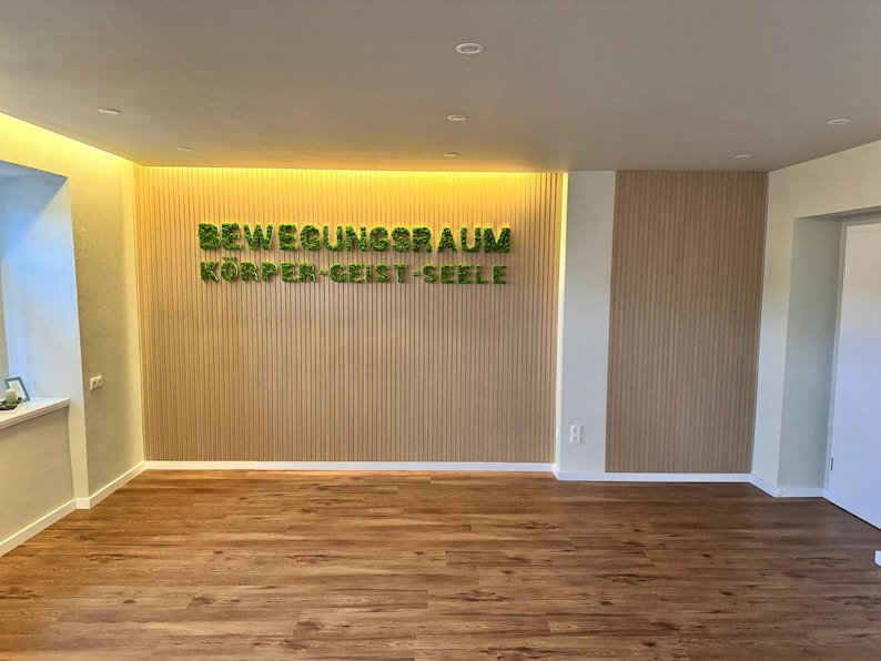 Pu&ograve; includere: Una stanza vuota con pavimento in legno e pareti chiare. La parete di fondo presenta pannelli di legno verticali con il testo verde "BEWEGUNGSRAUM K&Ouml;RPER-GEIST-SEELE". L'illuminazione a incasso illumina lo spazio, creando un'atmosfera luminosa e accogliente.