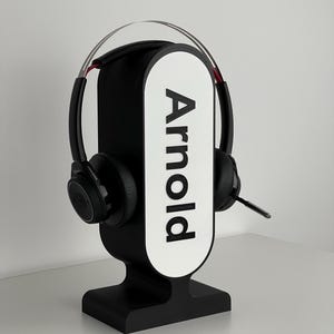 Könnte beinhalten: Schwarzer Headset-Ständer mit einem weißen ovalen Schild, auf dem "Arnold" steht. Das Headset ist schwarz mit einem silbernen Kopfbügel.