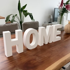 Op de afbeelding: Witte houten letters met de tekst "HOME" staan op een houten tafel. De letters zijn een opvallend decoratief element. De tafel staat in een kamer met planten en andere meubels, wat een uitnodigende sfeer creëert.